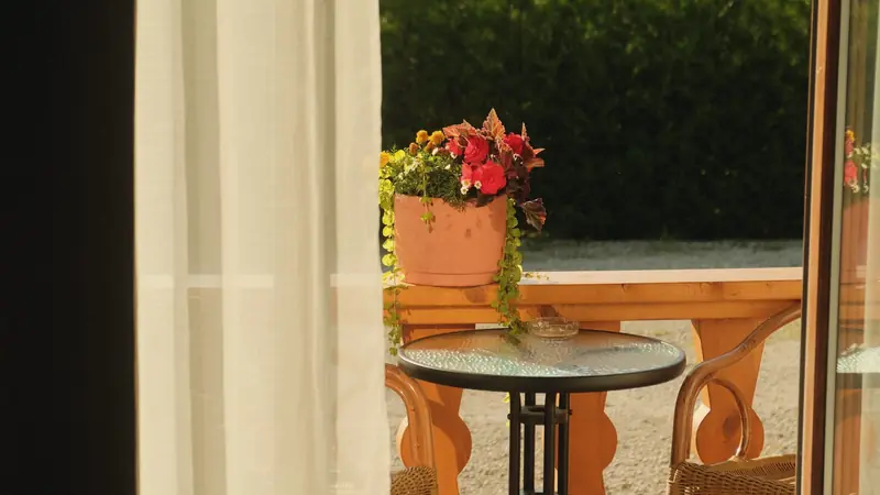 Baie vitrée ouverte sur une terrasse en bois, voilage blanc au premier plan, table ronde et fauteuils en osier au soleil du soir