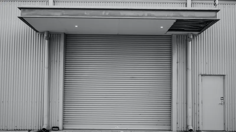 Porte enroulable à lames métalliques fermée sous un auvent, sur un bâtiment en bardage, photo en noir et blanc