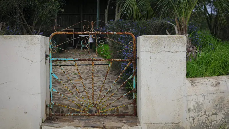 Petit portillon en fer forgé rouillé, peint en bleu turquoise à l'origine, fermé entre deux piliers maçonnés blancs, avec une allée pavée derrière